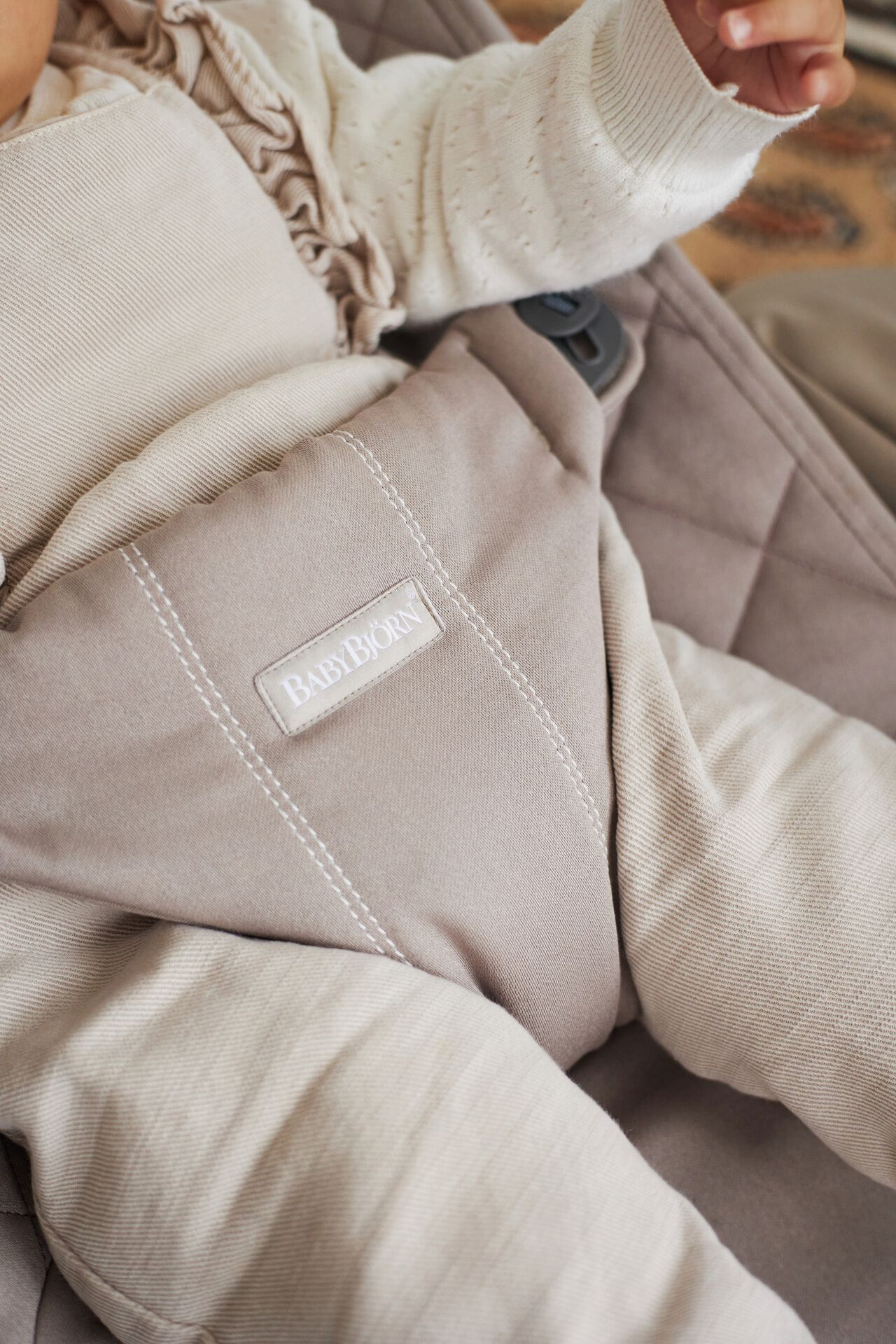 A baby wearing a beige onesie is seated in a beige BabyBjörn carrier. The focus is on the carrier's logo and the baby's legs.