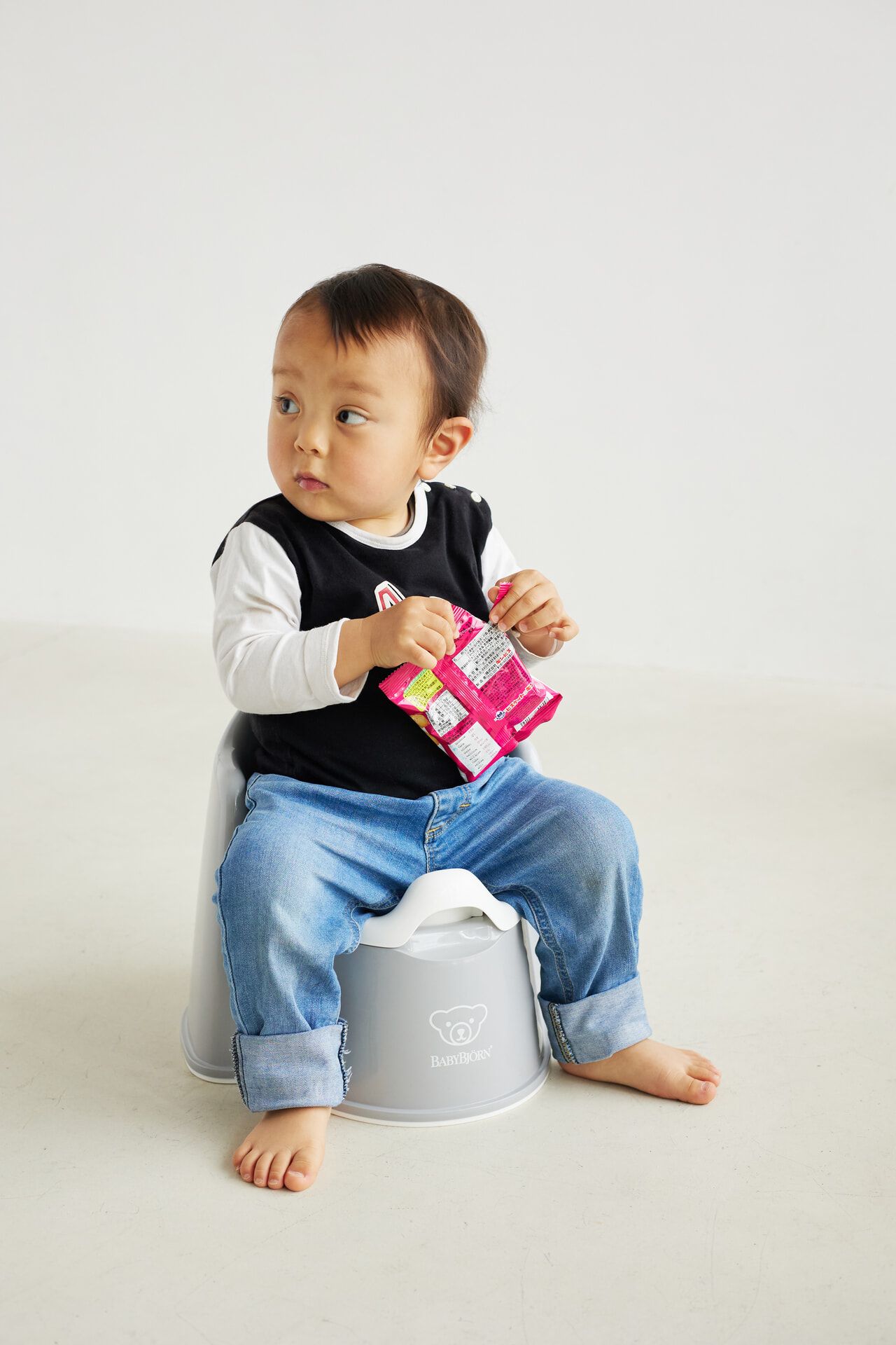 A toddler sits on a gray potty, holding a colorful snack package, wearing a black and white shirt with blue jeans, looking to the side.
