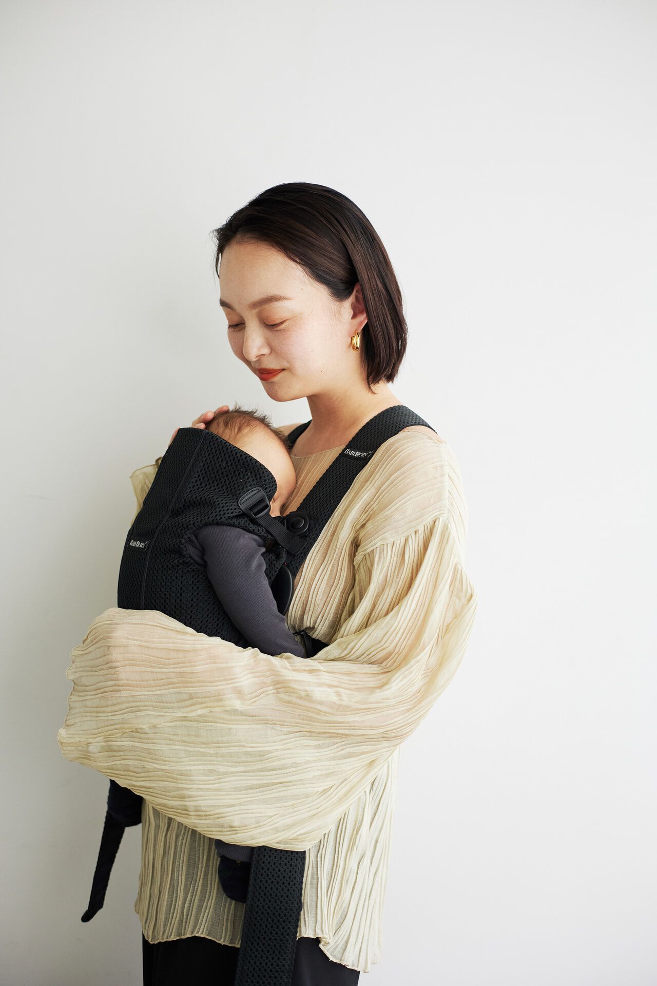 A woman gently holds a baby in a black carrier against her chest, wearing a light, textured blouse, standing against a plain white background.