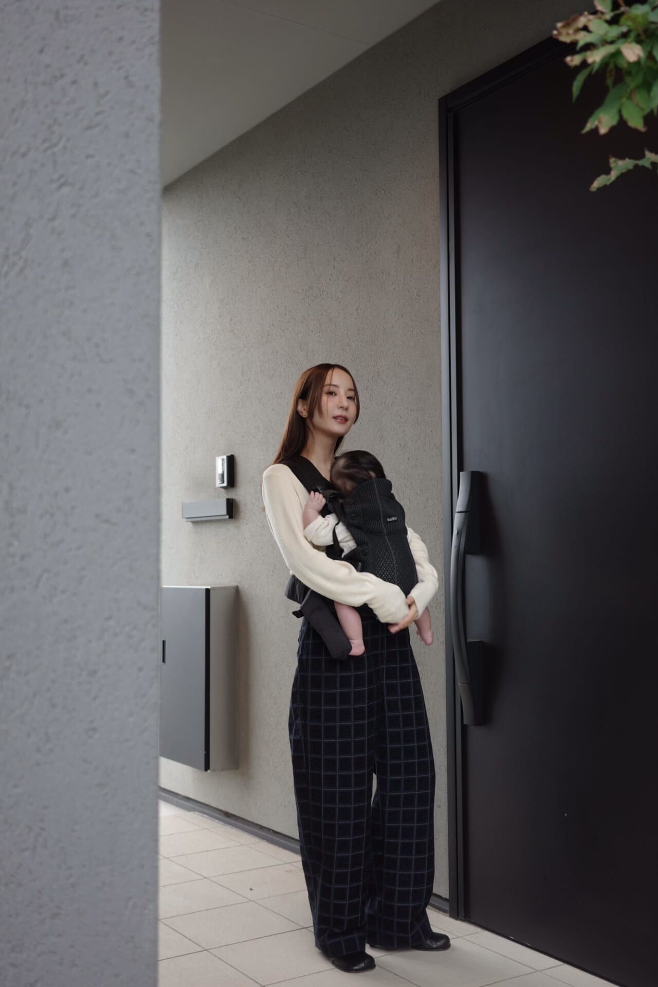 Woman standing by a modern door, wearing a cream sweater and checkered pants, carrying a baby in a black carrier.