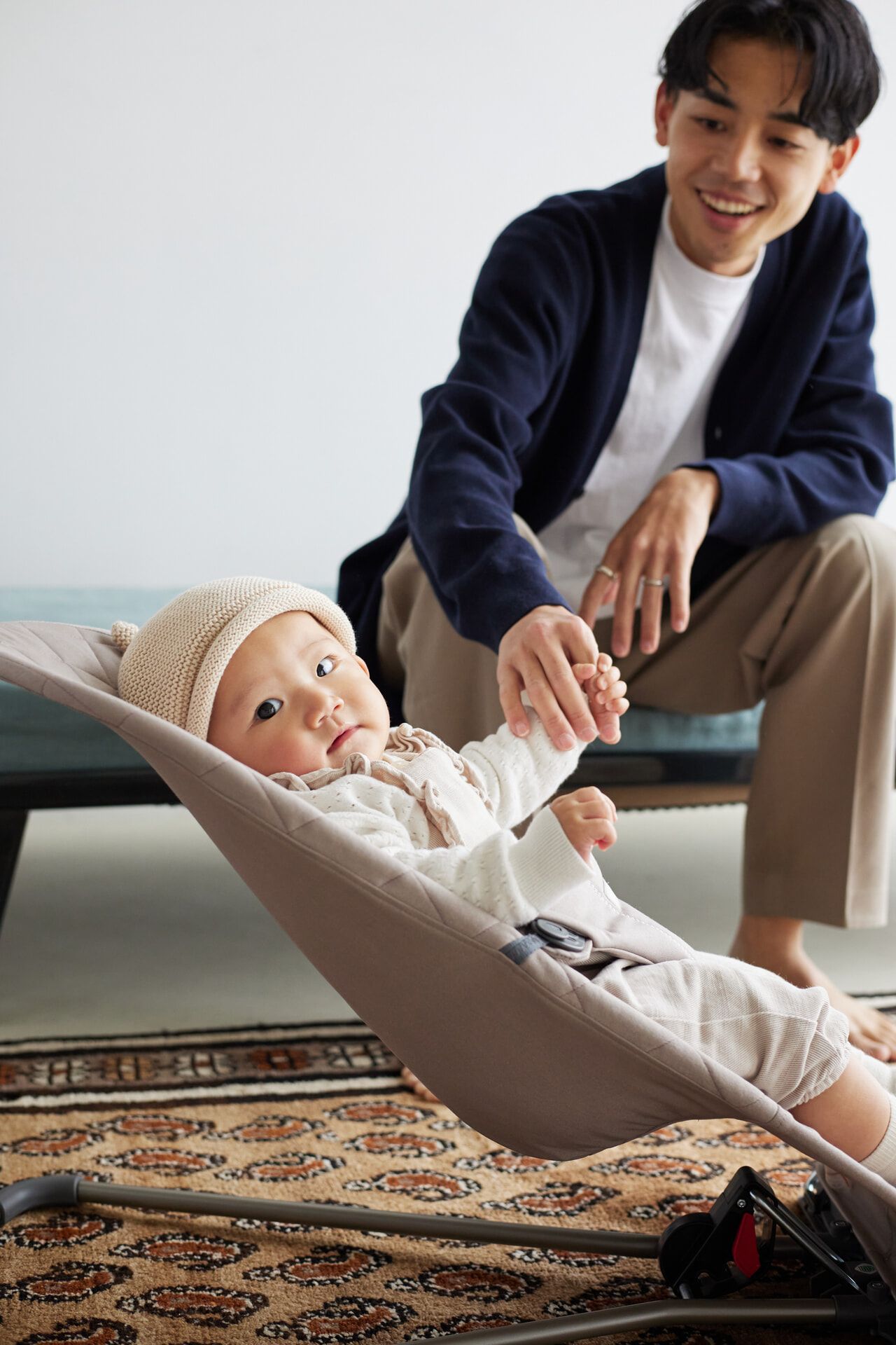 A baby in a beige outfit and hat sits in a bouncer, reaching for an adult's hand. The adult, sitting nearby, smiles at the baby.