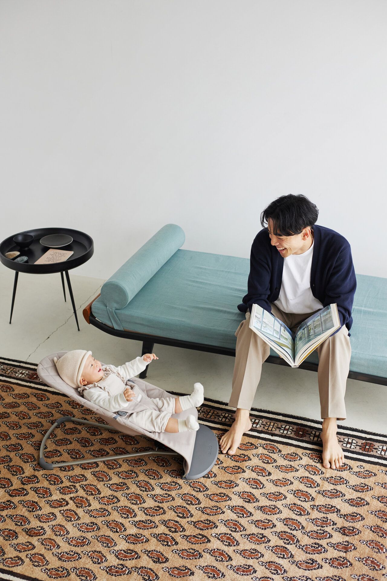 A man sits on a sofa reading a newspaper, smiling at a baby in a bouncer. The room has a patterned rug and a small round table.