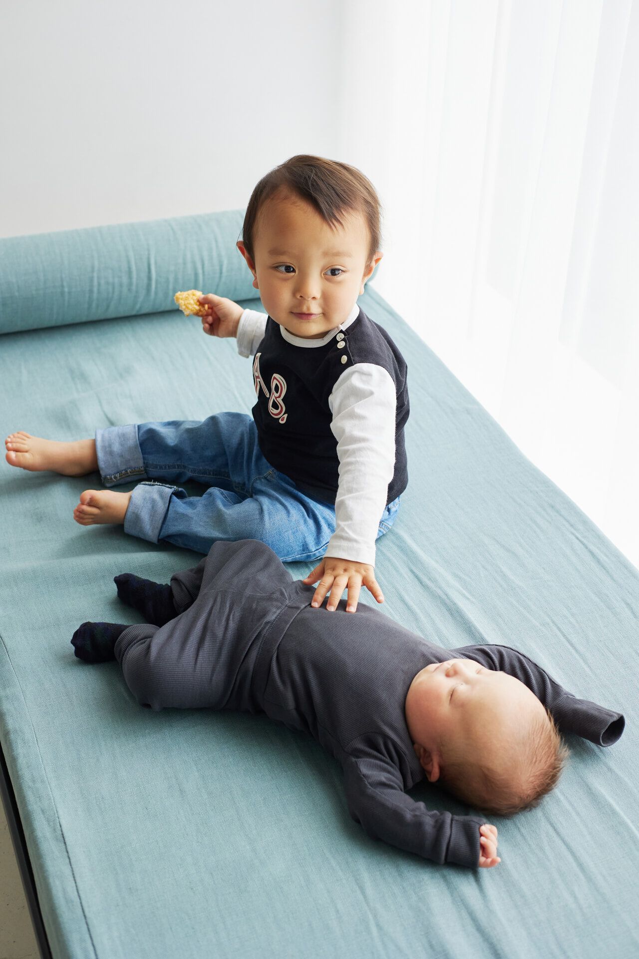 A toddler sits on a teal couch holding a cracker, gently touching a sleeping baby lying beside them.