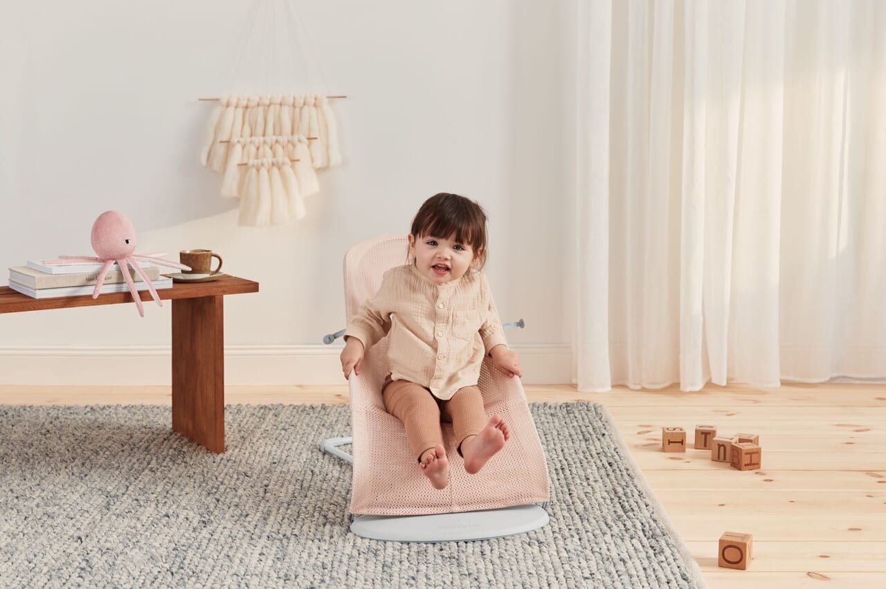 A baby sits in a pink bouncer on a light wooden floor, surrounded by toys and a small table, with curtains and a textured rug in the background.