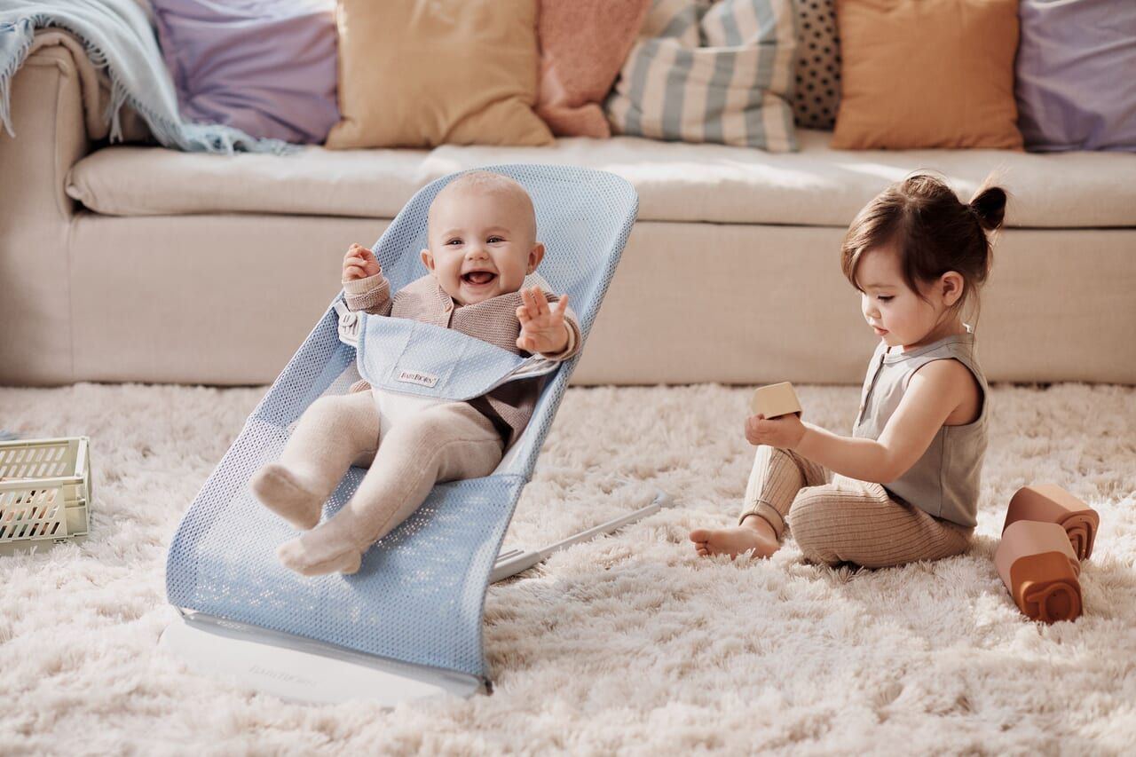 A baby in a blue bouncer smiles while a toddler plays with blocks on a fluffy rug in a cozy living room.