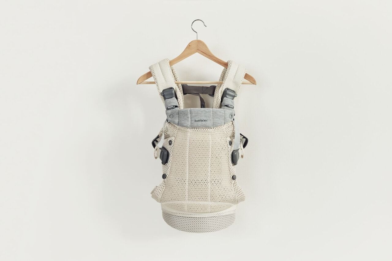 A beige baby carrier with adjustable straps and buckles hangs on a wooden hanger against a plain white background.