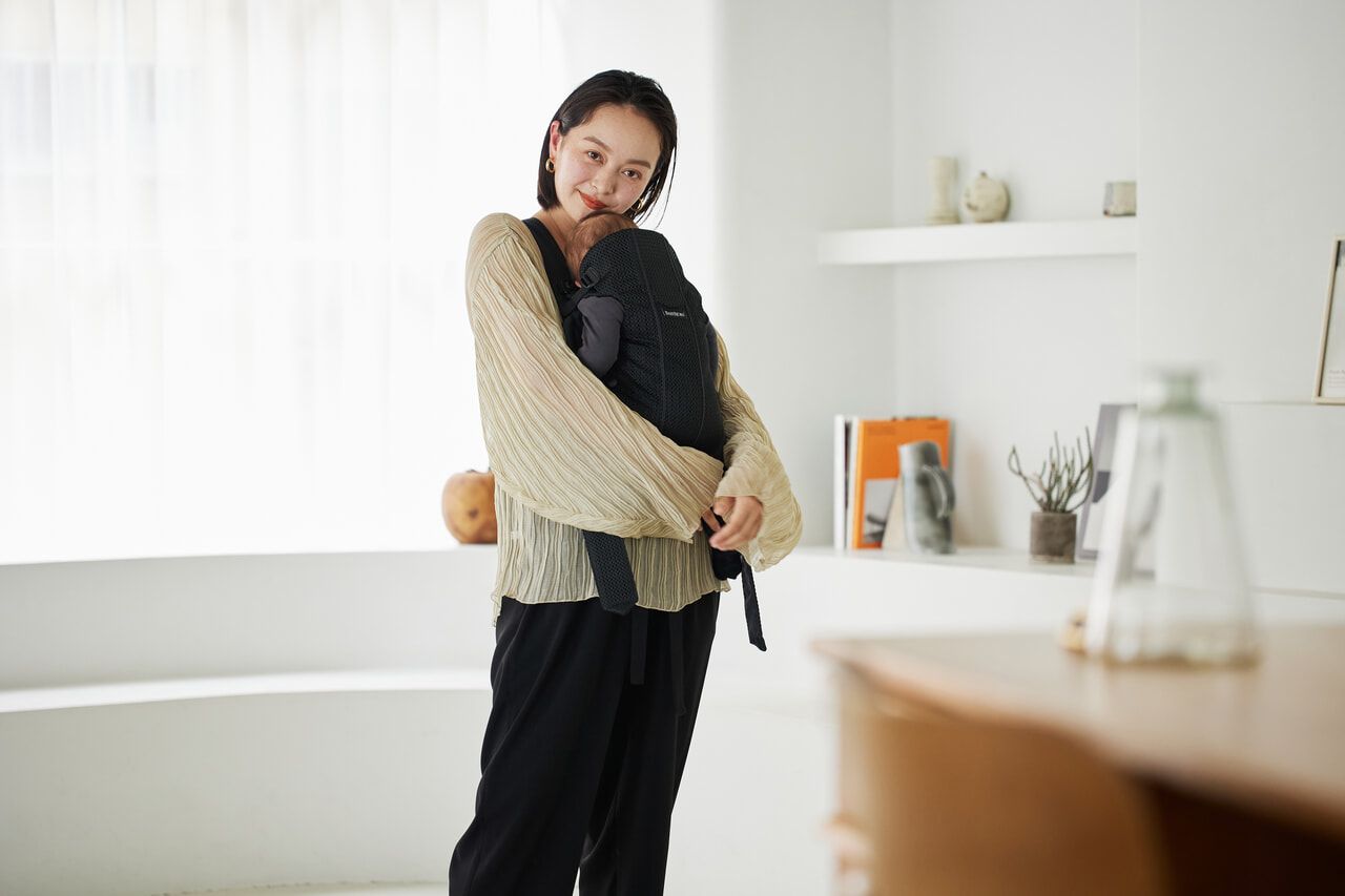 A woman in a cozy room holds a baby in a black carrier. She wears a beige sweater and black pants, standing near a window with soft lighting.