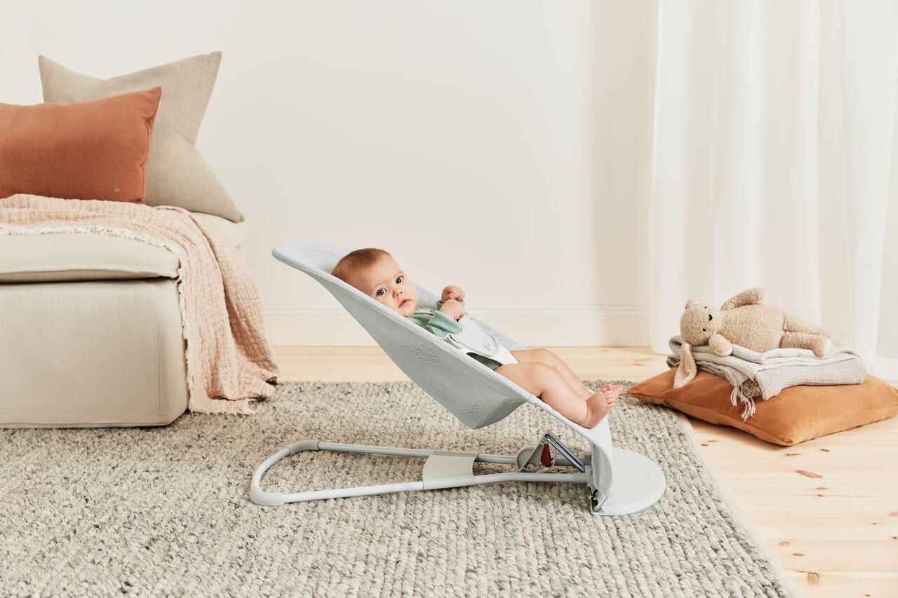 Baby in a gray bouncer on a beige rug, with cushions and a teddy bear on a pillow nearby in a cozy living room setting.