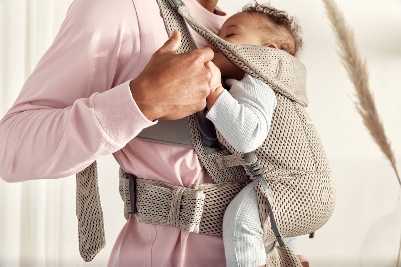 Person in a pink shirt carrying a baby in a beige mesh baby carrier. The baby is wearing a light blue outfit and appears to be sleeping.