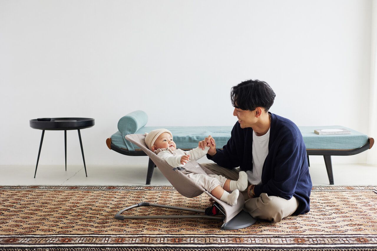A man kneels on a patterned rug, smiling at a baby in a bouncer. The room has a minimalist design with a bench and a small round table.
