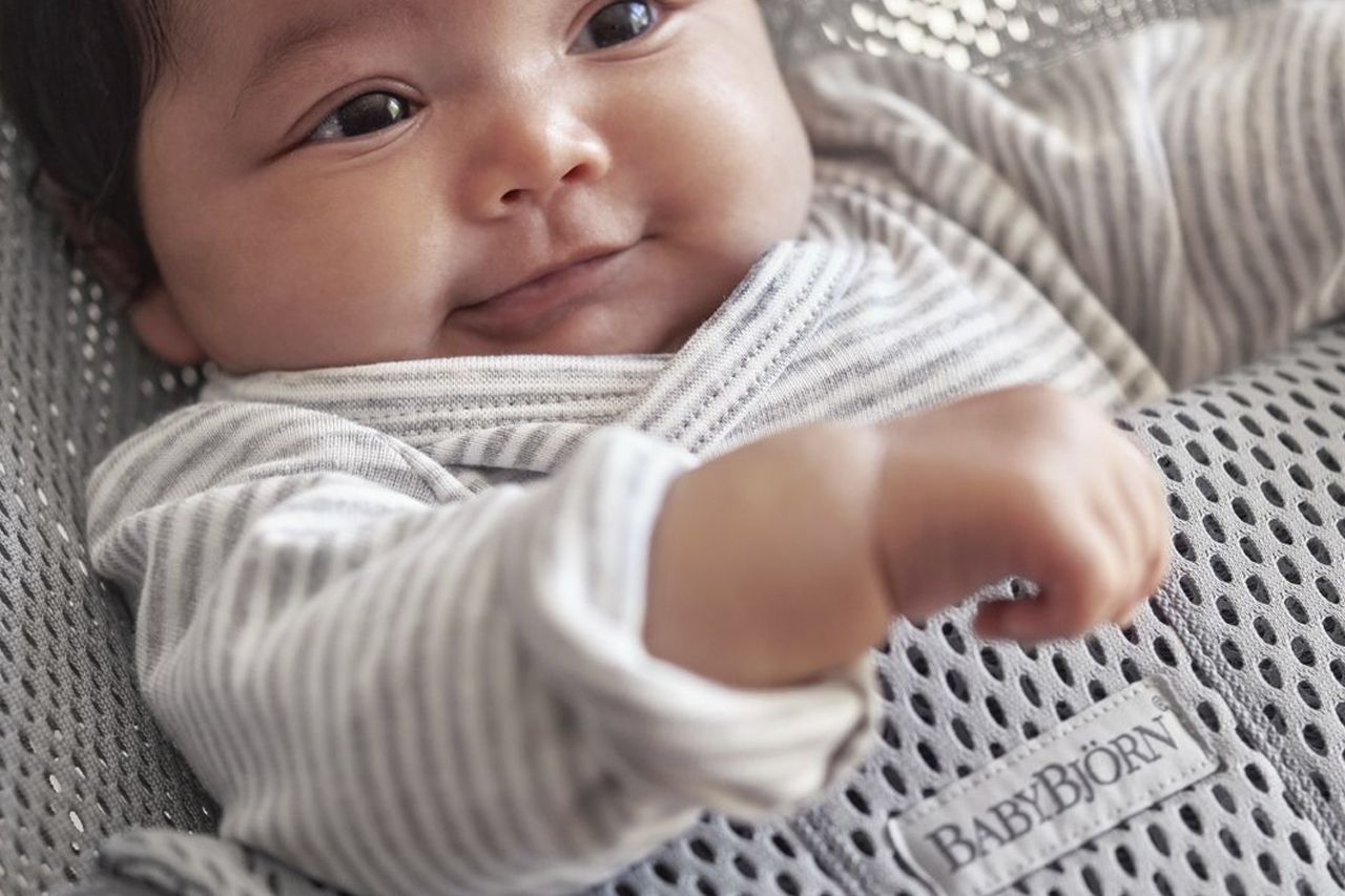 Smiling baby in a striped outfit lying on a gray BabyBjörn bouncer.