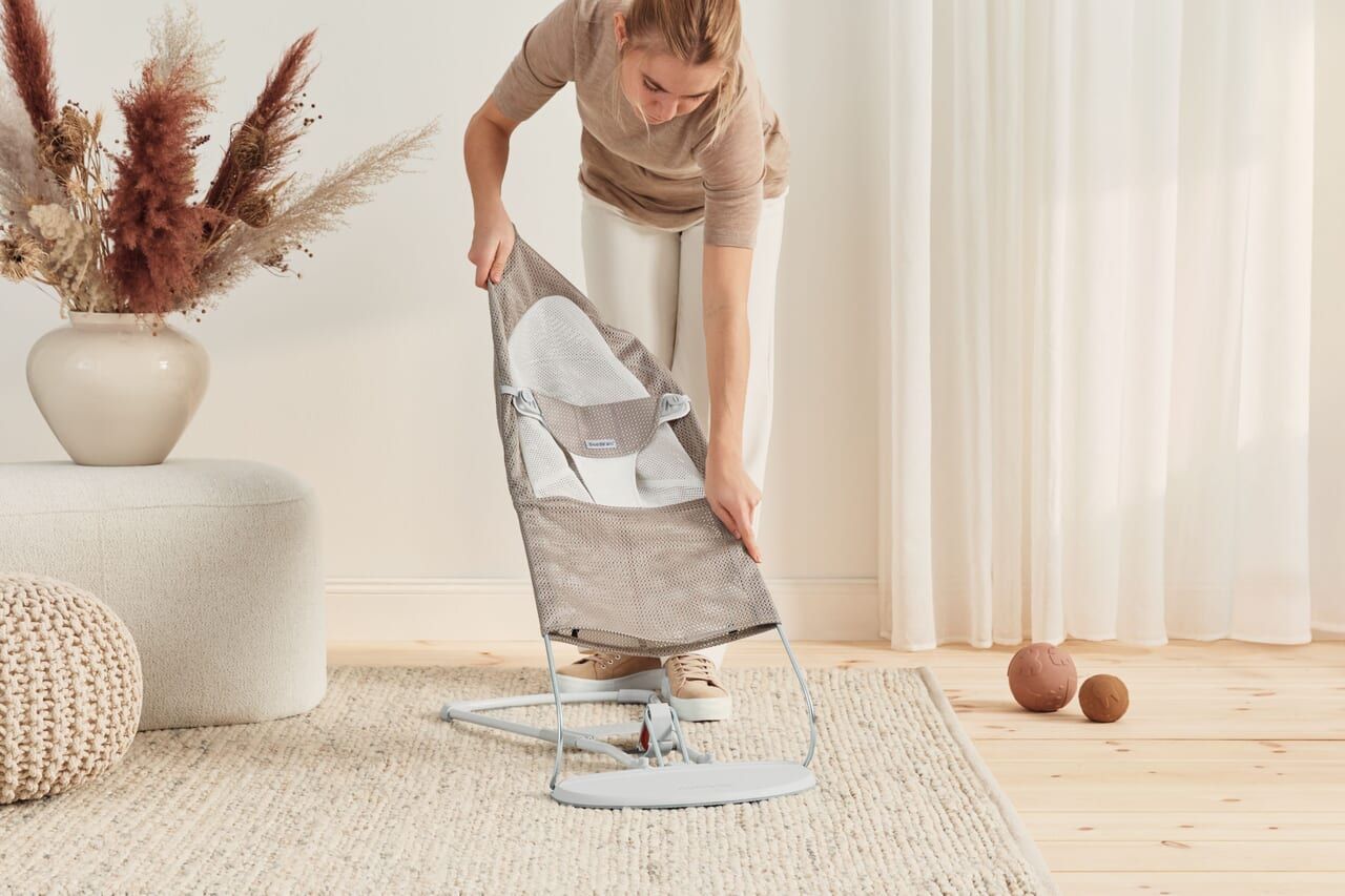 A woman adjusts a baby bouncer in a cozy living room with a soft rug, pouf, and decorative vase with dried flowers.