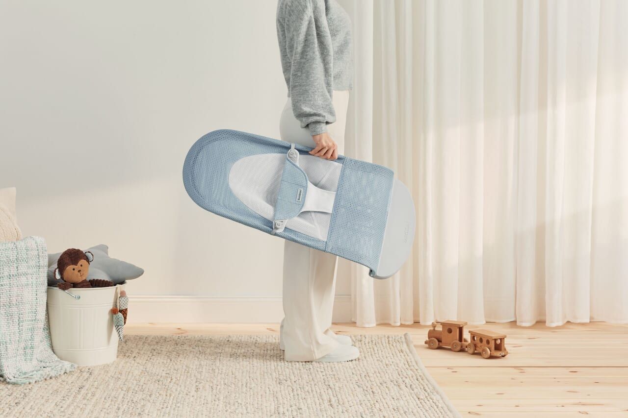 Person holding a folded blue baby bouncer in a bright room with a toy basket and wooden train on the floor.