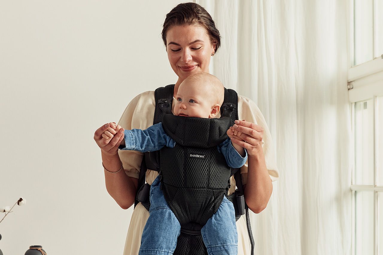 A person smiling while holding a baby in a front-facing carrier indoors. The baby is wearing a blue outfit.