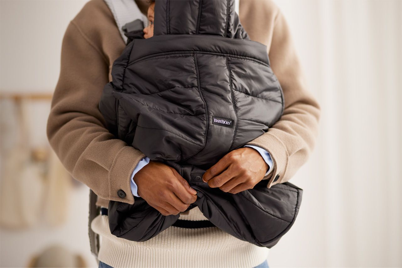 Person wearing a beige coat adjusts a black padded baby carrier with a baby inside, in a softly lit room.