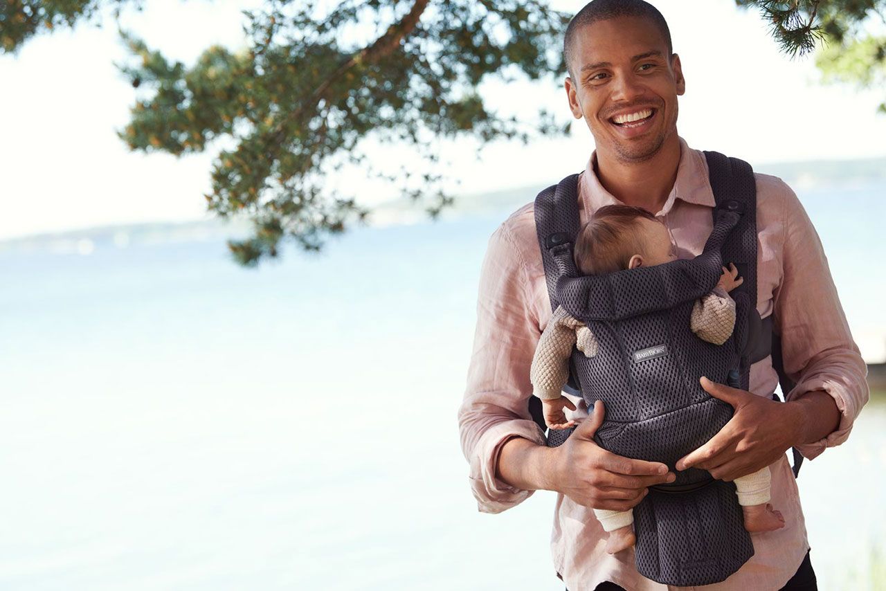 Smiling man holding a baby in a carrier by a lake, with trees in the background.