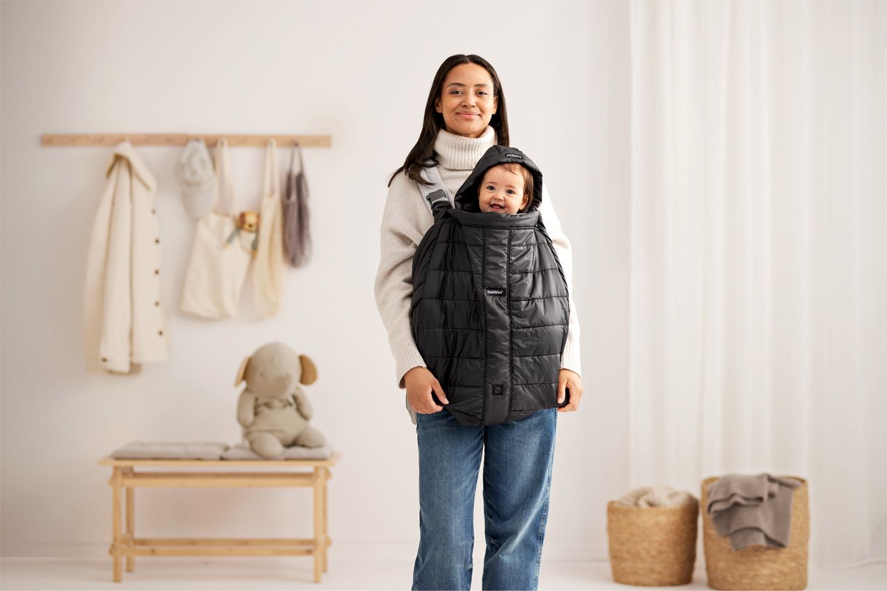 Woman smiling with a baby in a black carrier, standing in a cozy, neutral-toned room with a bench, hanging clothes, and baskets.