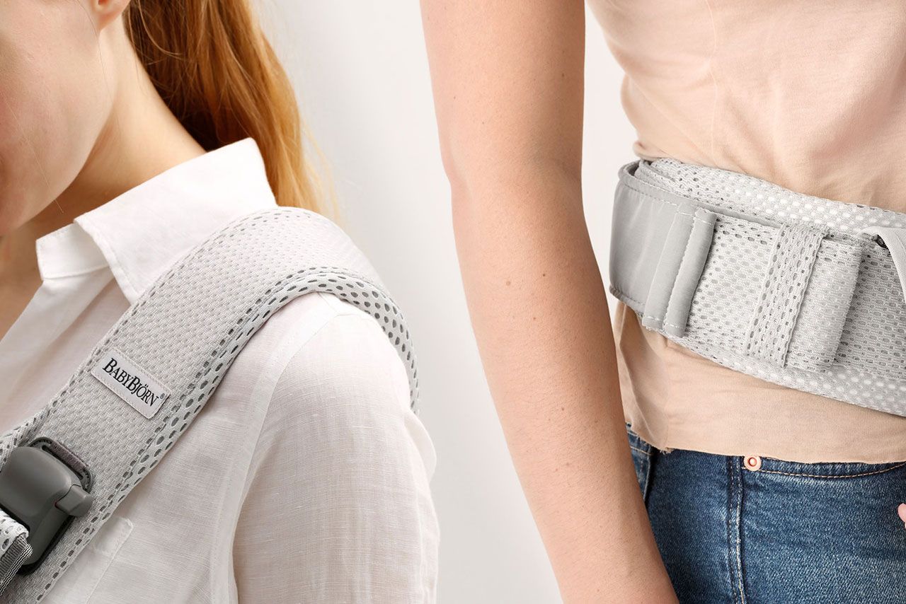 Close-up of two people wearing a gray BabyBjörn carrier. One shows the shoulder strap, the other the waist belt. Both wear casual clothes.