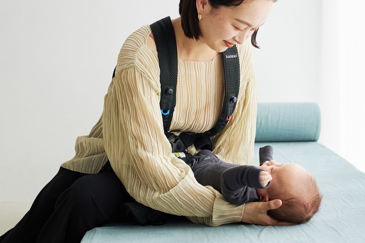 A woman wearing a beige blouse and black harness cradles a baby on a light blue cushion, looking down affectionately.