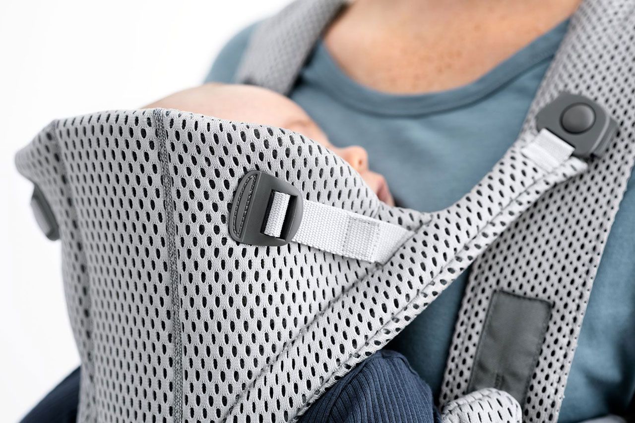 Close-up of a baby sleeping in a gray, breathable mesh baby carrier worn by an adult in a blue shirt.