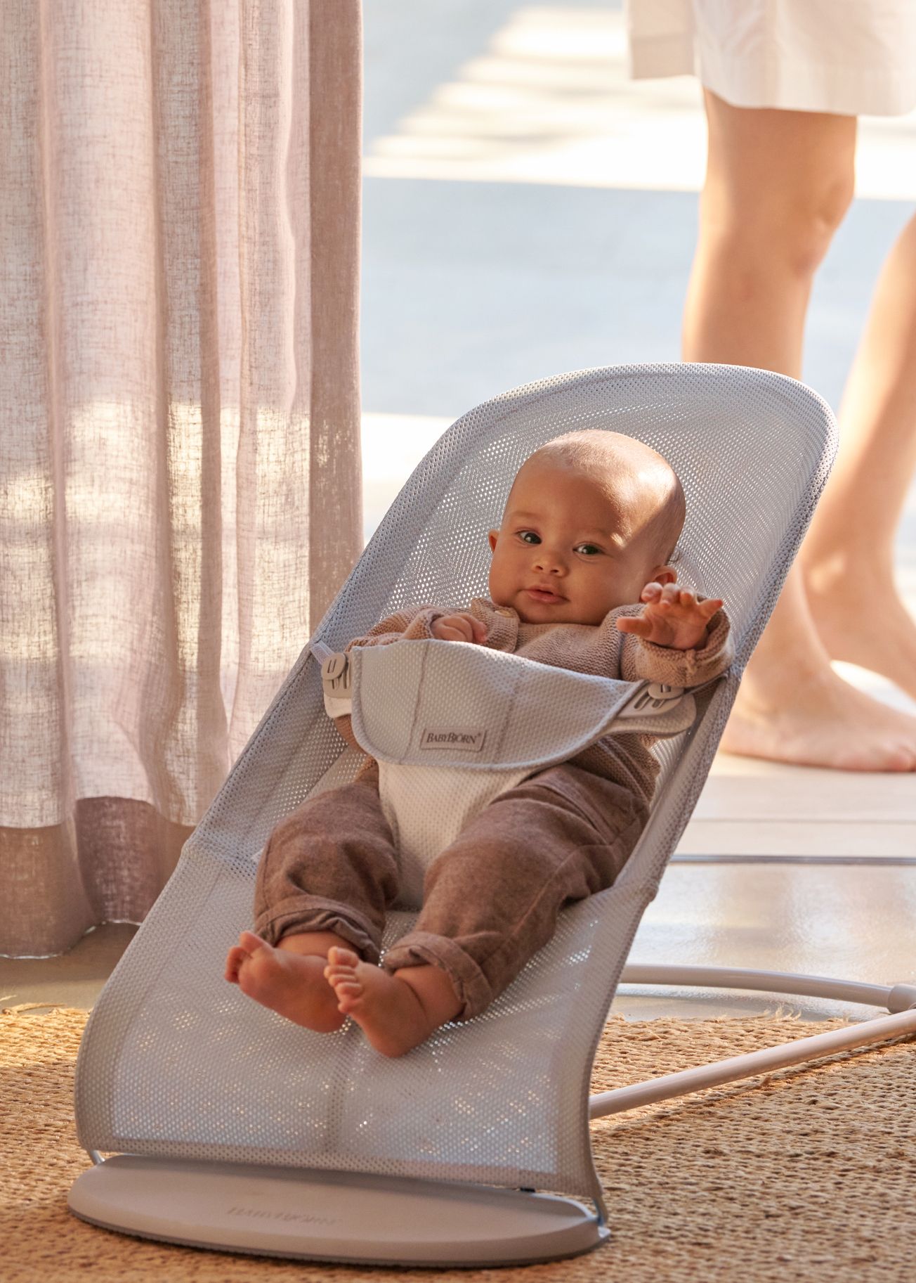 Baby sitting in a silver white mesh baby bouncer, wearing a brown outfit. Sunlight filters through curtains, and an adult's legs are visible in the background.