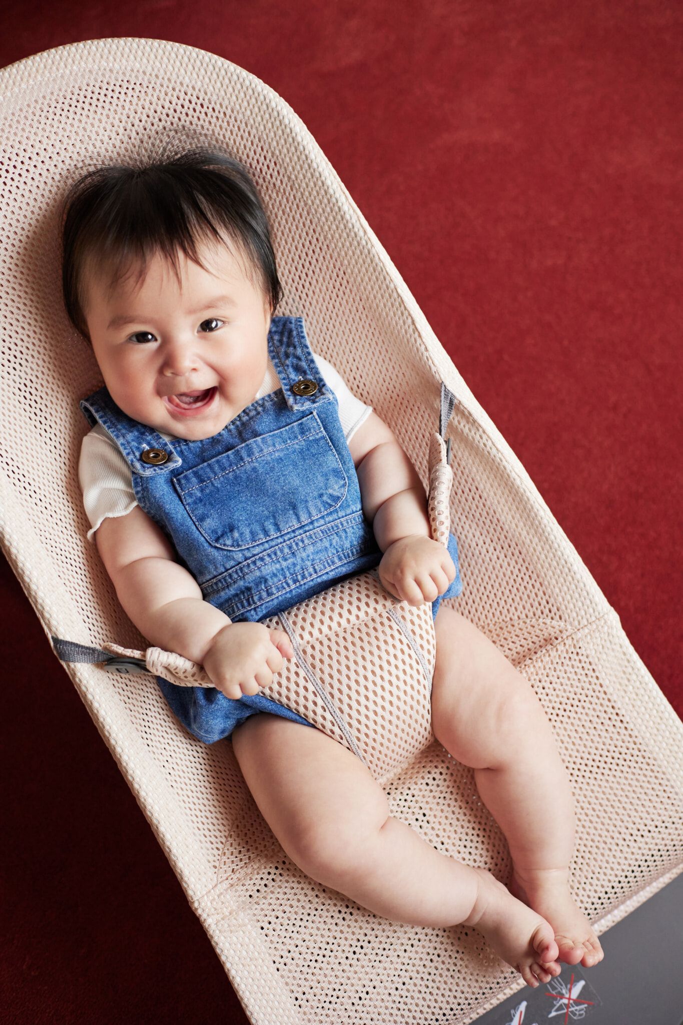 Smiling baby in denim overalls sits in a mesh baby bouncer on a red carpet.