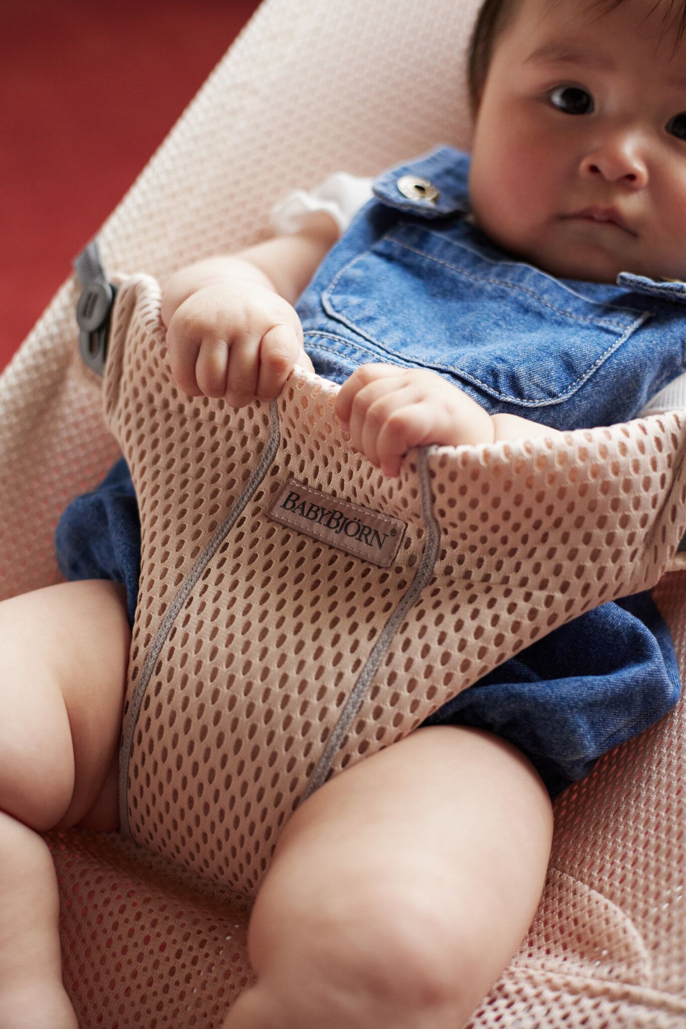 Baby in denim overalls sits in a beige mesh BabyBjörn bouncer, holding the fabric, with a focused expression.