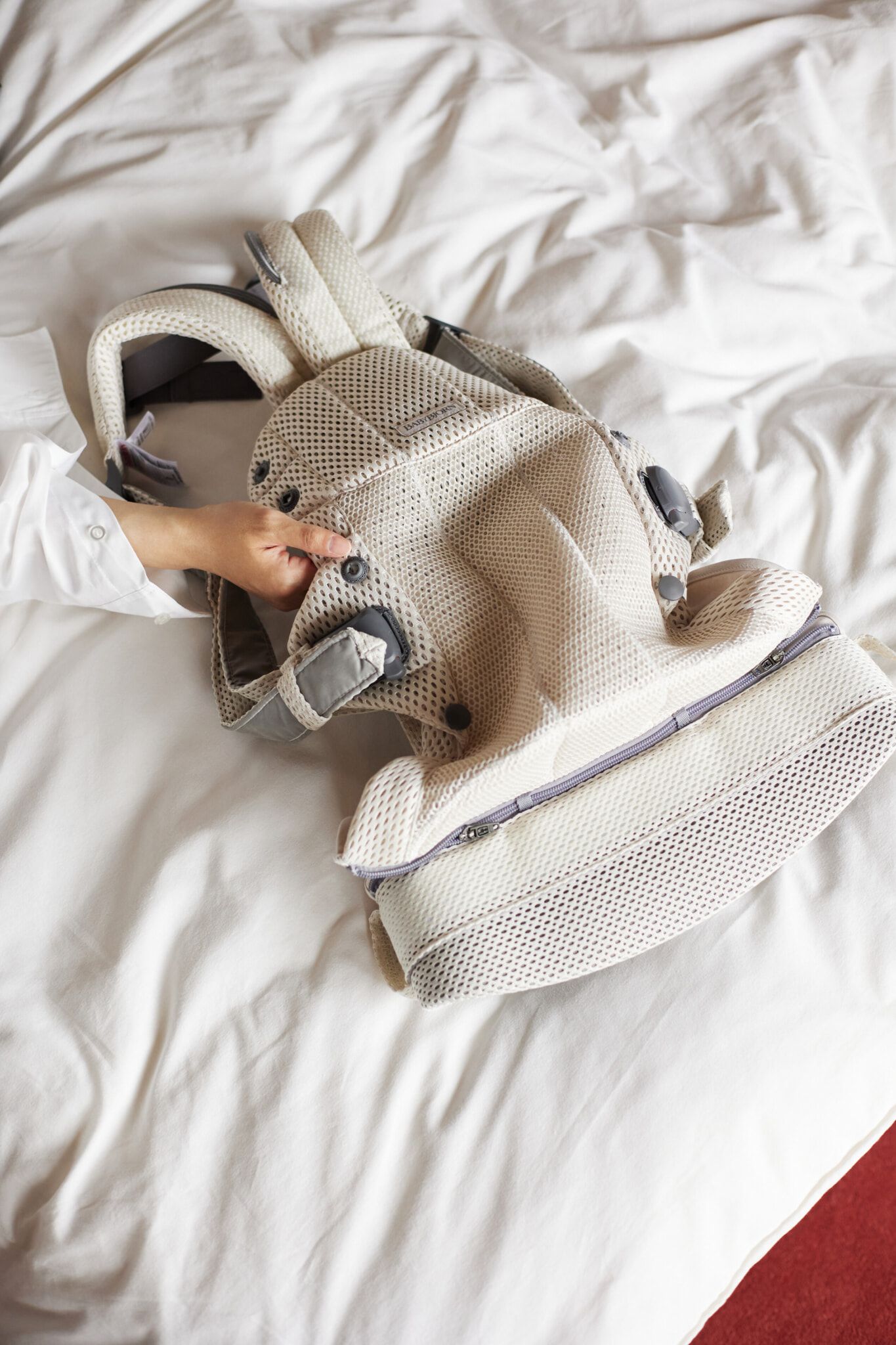 A person adjusts a beige mesh baby carrier with straps, laid out on a white bedspread.