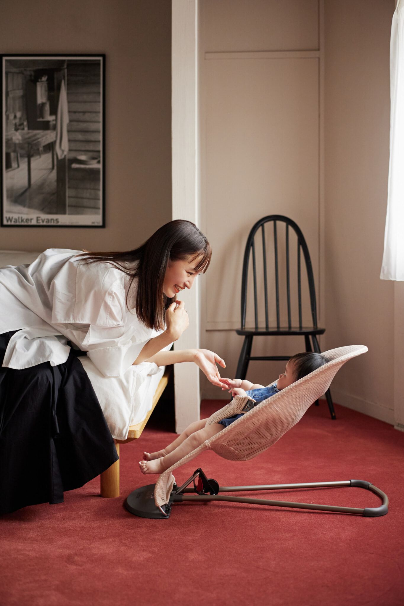 A woman gently interacts with a baby in a bouncer on a red carpeted floor, in a room with a poster and a black chair.