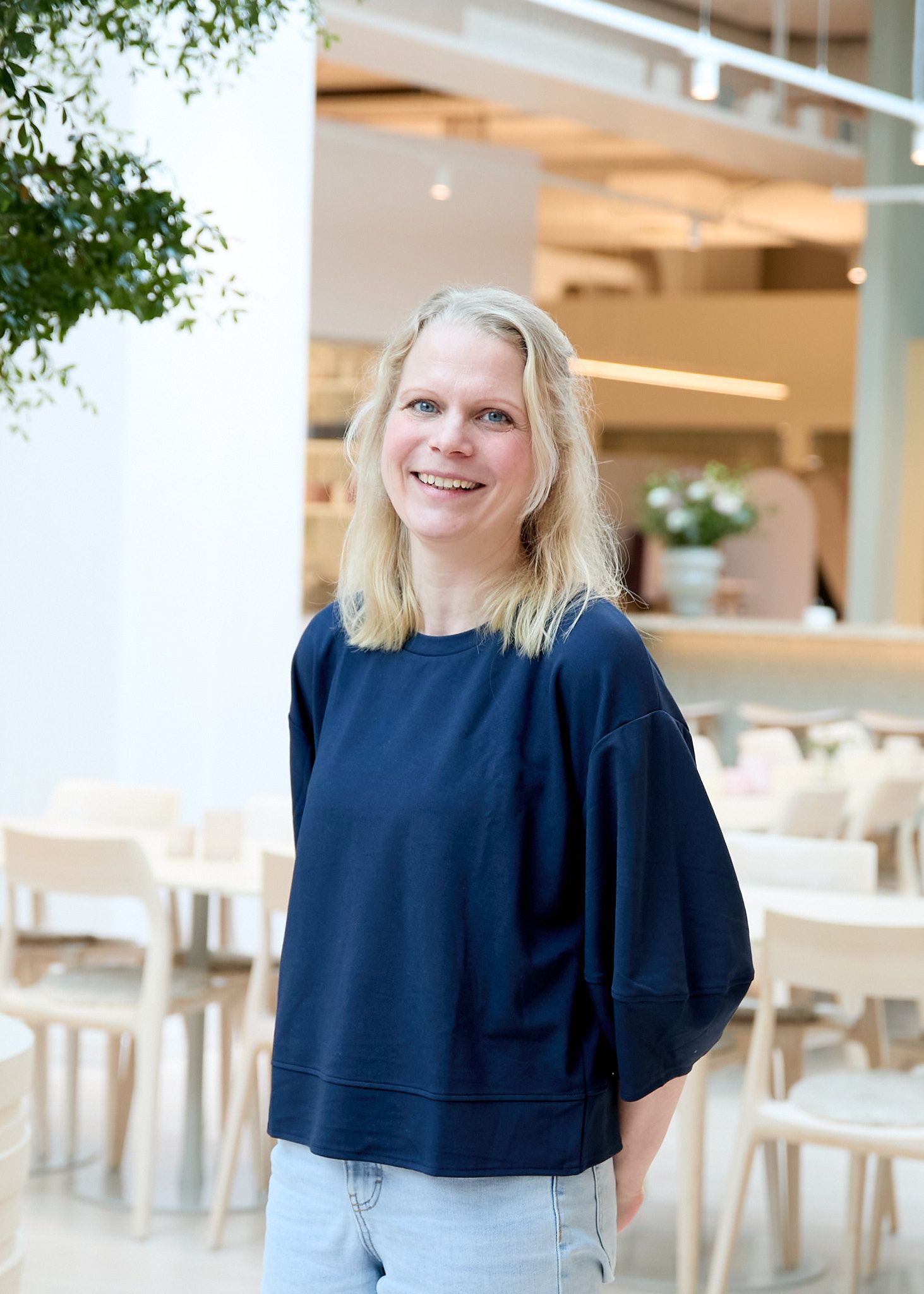 Smiling person with blonde hair in a blue shirt, standing in a modern, well-lit indoor space with light wooden furniture and a plant.