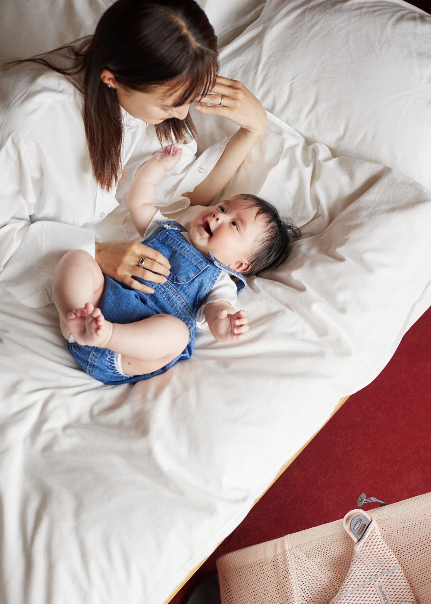 A woman in a white shirt lovingly gazes at a baby in denim overalls, lying on a white bed with a red floor visible.