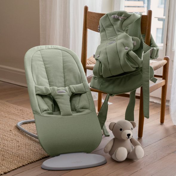 A green baby bouncer and carrier sit on a wooden chair. A beige teddy bear is on the floor nearby, in a softly lit room.