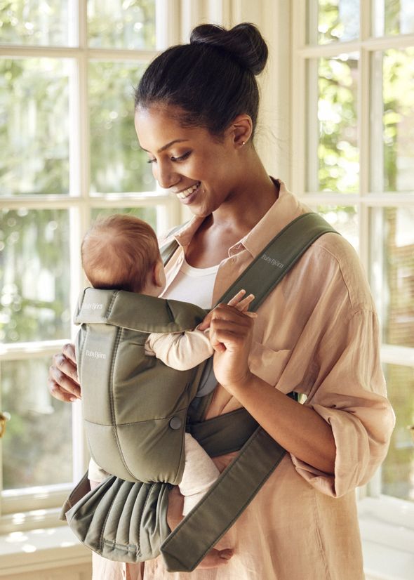 A woman smiles while holding a baby in a green carrier near a window.