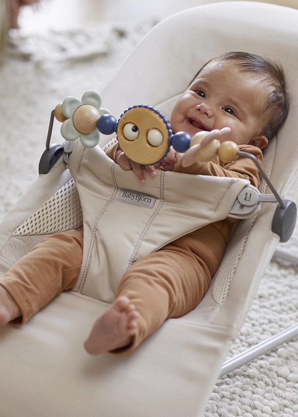 Smiling baby in a beige bouncer playing with a colorful toy bar. The baby wears a brown outfit and is sitting indoors on a textured rug.