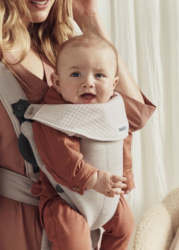 A happy baby in a brown outfit is in a white baby carrier, worn by an adult, smiling at the camera in a softly lit room.