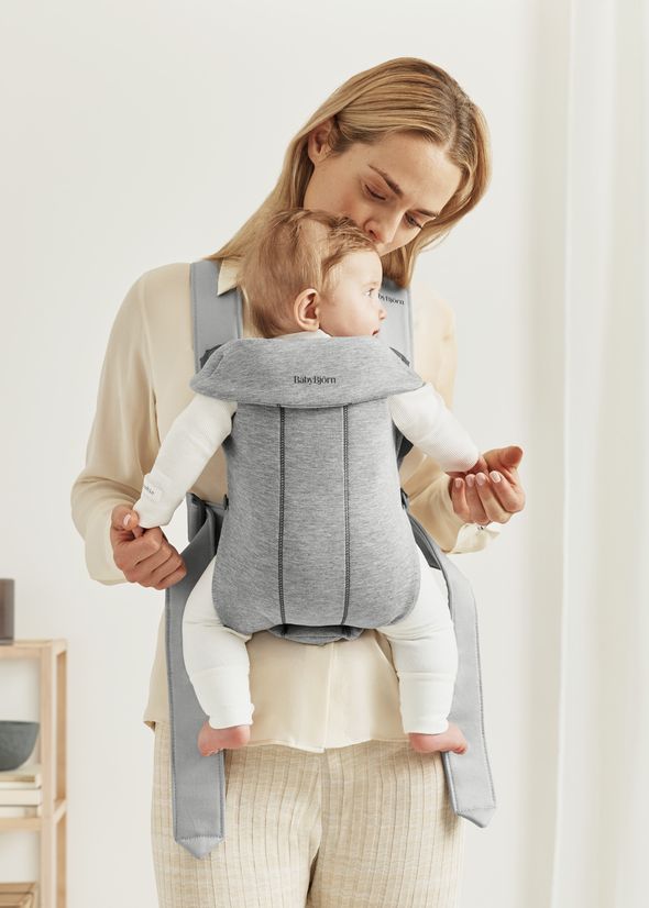 A woman wearing a white blouse holds a baby in a gray baby carrier, smiling as she looks at the baby. The setting is a bright room.