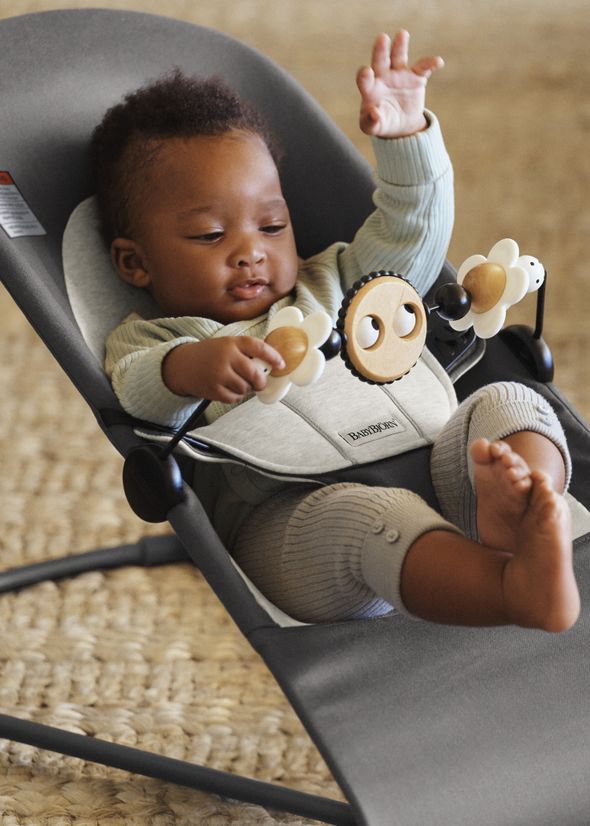 Baby in a gray bouncer, wearing a light outfit, playing with attached toys, and reaching up with one hand.
