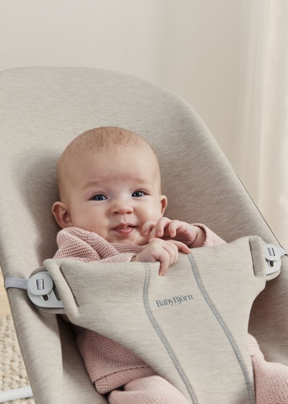 A baby in a pink outfit sits comfortably in a beige BabyBjörn bouncer, smiling and holding their hands together.