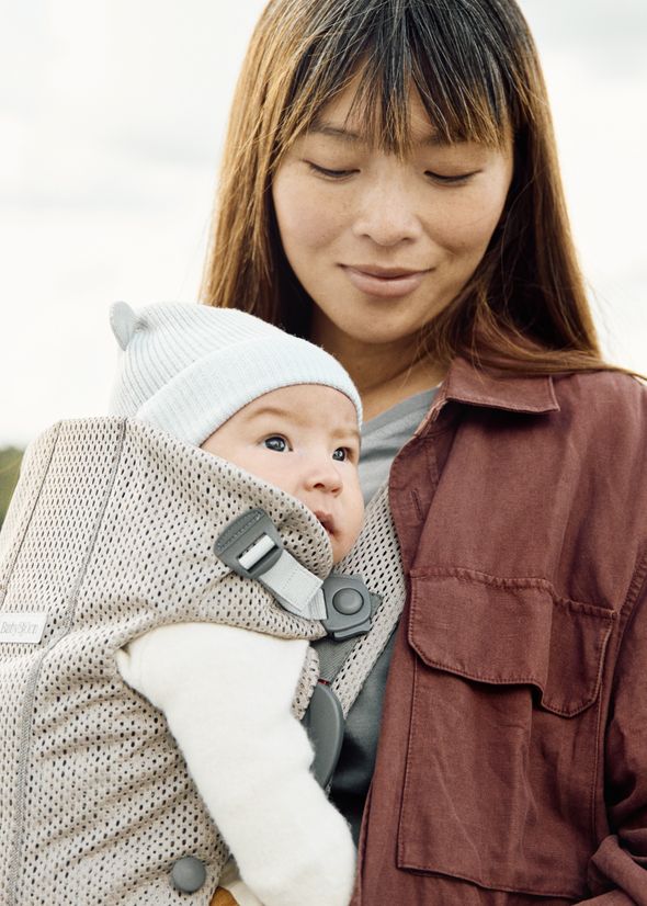 A woman smiles gently at a baby in a front carrier. The baby wears a light blue beanie and looks curiously to the side.