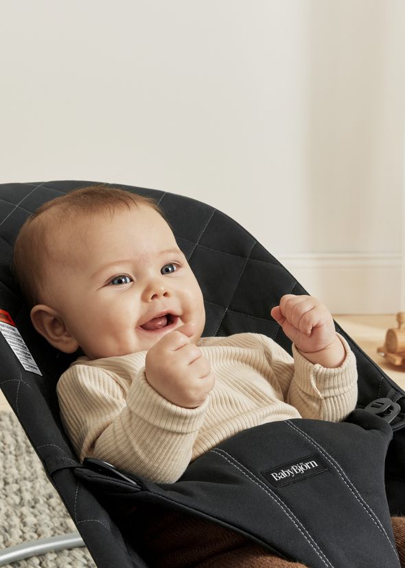 A happy baby in a beige top sits in a black BabyBjörn bouncer, smiling and raising both fists, with toys in the blurred background.