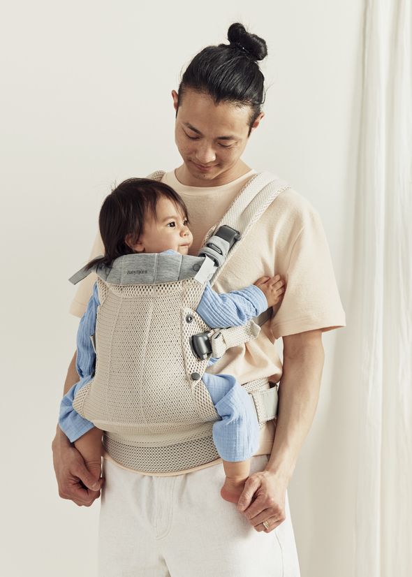 A man in a beige shirt holds a baby wrapped in a light-colored carrier. The baby wears a blue outfit and looks to the side.