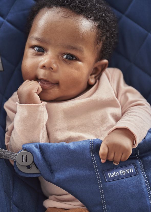 A baby in a pink outfit sits in a blue BabyBjörn seat, looking up with a hand near their mouth and a curious expression.