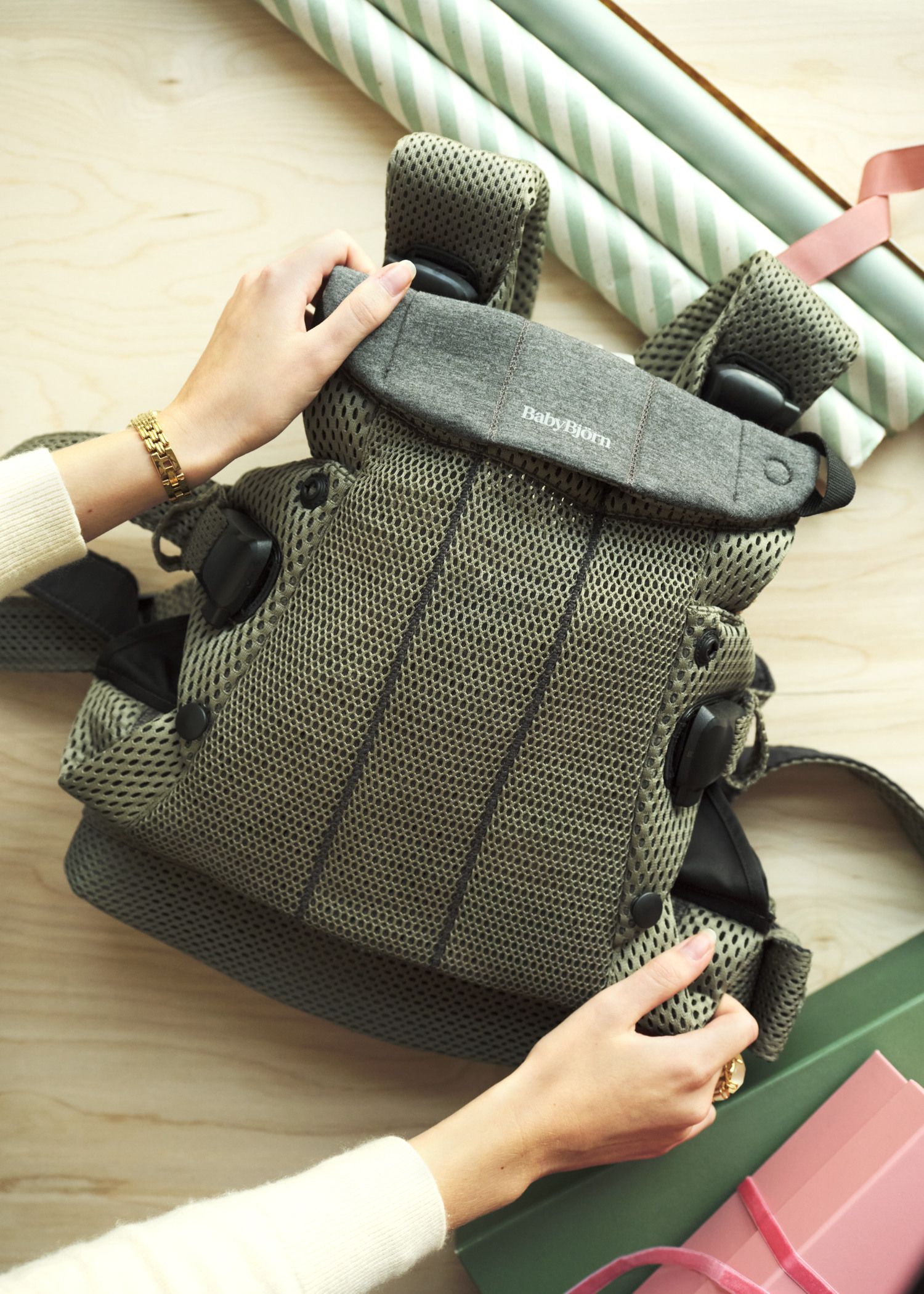 Hands holding a gray mesh baby carrier on a wooden surface, surrounded by gift wrapping paper and ribbons.