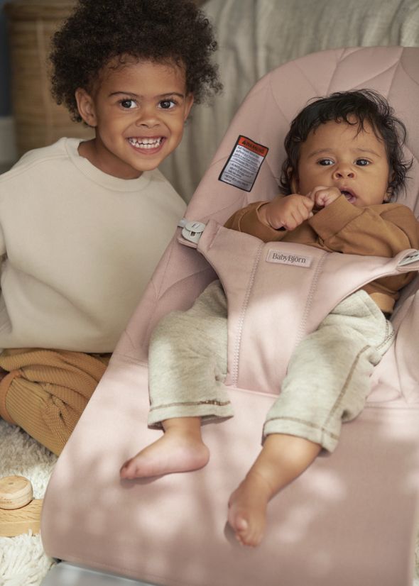 Two young children, one sitting and smiling, and the other lying in a baby bouncer. Both are wearing cozy, neutral-toned clothing.