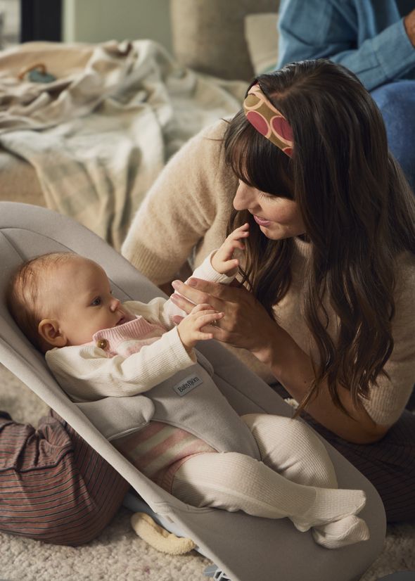 Woman gently holds a baby's hand as the infant lies in a gray bouncer. The woman smiles, wearing a headband. Cozy setting with a blanket nearby.
