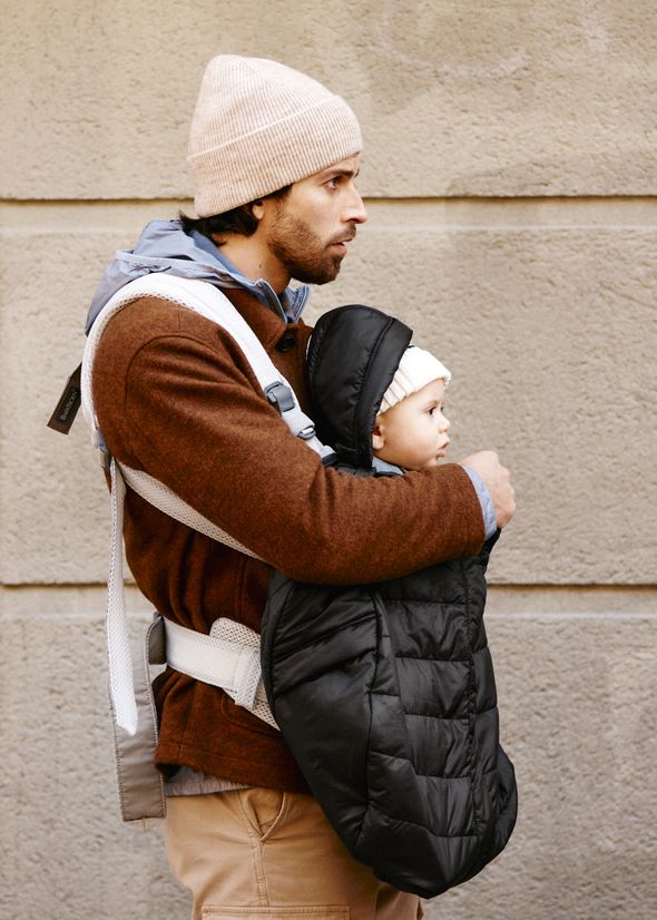 Man wearing a brown sweater and beanie carries a baby in a black carrier, both dressed warmly, walking along a textured wall.