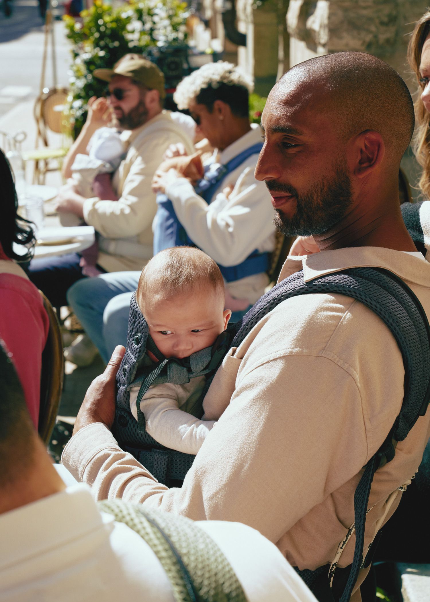 A man holds a baby in a carrier, surrounded by people sitting at an outdoor cafe. Sunlight casts shadows on the scene.