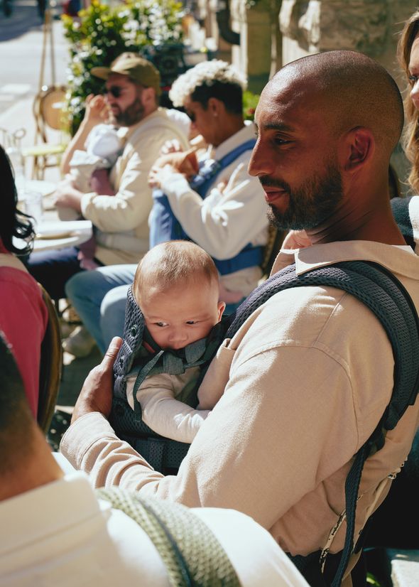 A man holds a baby in a carrier, surrounded by people sitting at an outdoor cafe. Sunlight casts shadows on the scene.