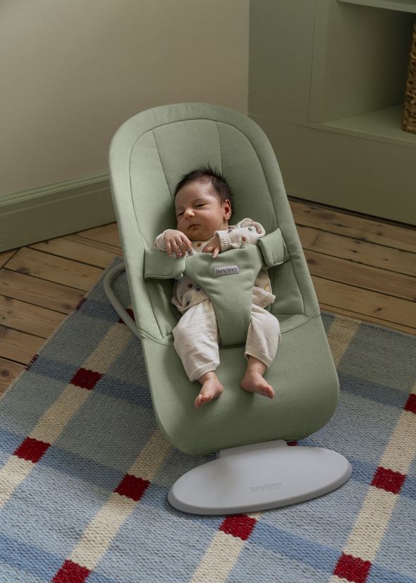 A baby sleeping peacefully in a green bouncer on a checkered rug next to a wooden floor.