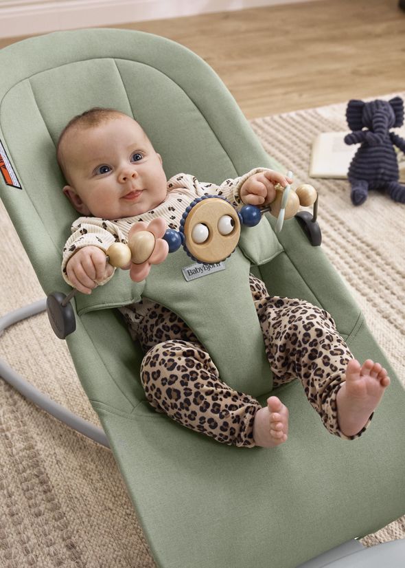 Baby in a green bouncer with leopard print pants, playing with a toy. A plush elephant and book lie on the floor nearby.